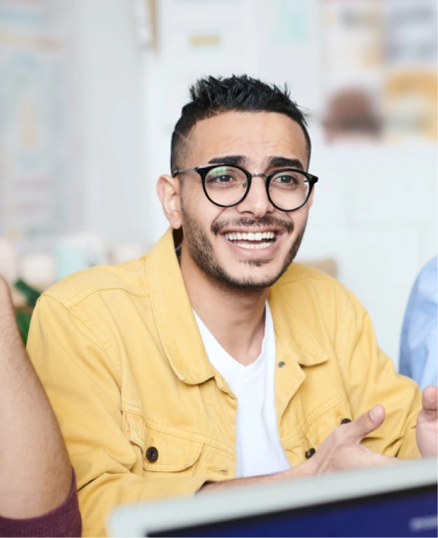 A smiling young man in glasses and a yellow jacket engaging in a lively team discussion.