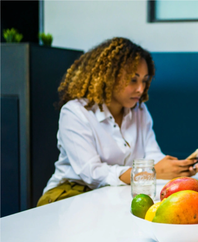 A professional woman with curly hair working on her phone at a bright communal table with fresh fruit.