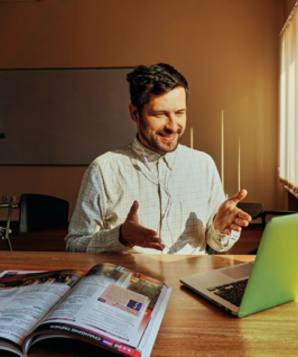 Un homme en visioconférence sur son ordinateur portable, faisant des gestes explicatifs dans un bureau ensoleillé.