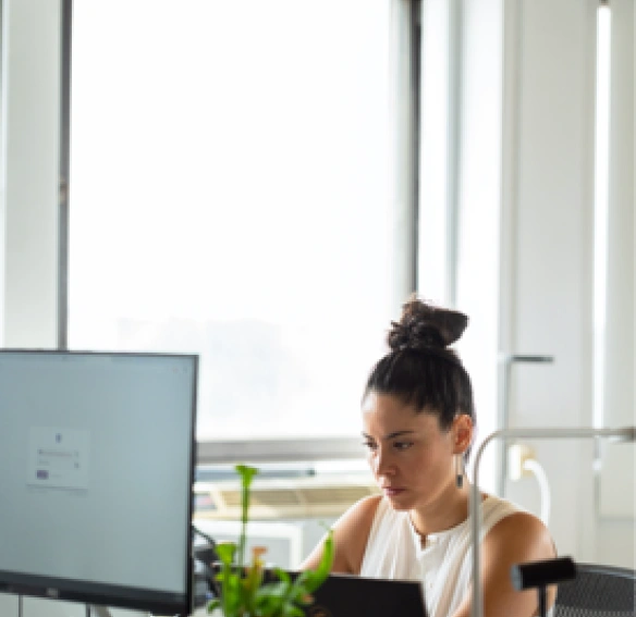 Une femme concentrée travaillant à son bureau avec plusieurs écrans dans un environnement professionnel moderne.