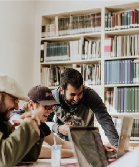Trois jeunes professionnels riant et collaborant devant un ordinateur dans une bibliothèque ou un espace de travail.