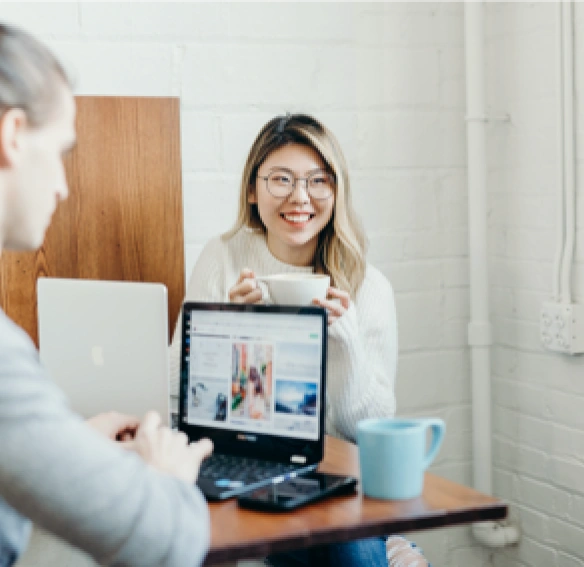 Deux collègues souriants discutant autour d'un café avec un ordinateur portable dans un bureau lumineux.