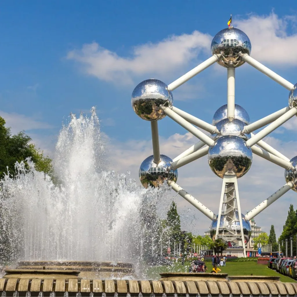 L'imposant monument de l'Atomium à Bruxelles se dresse sur un ciel bleu, encadré par une fontaine jaillissante et des arbres verdoyants, une icône de visibilité souvent optimisée par une agence seo belgique.