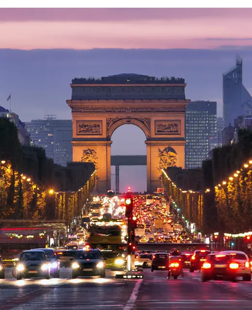 L'Arc de Triomphe illuminé, un monument que l'on pourrait retrouver mis en avant par une agence SEO France, domine la perspective depuis les Champs-Élysées au crépuscule, avec un trafic dense et des lampadaires créant une scène urbaine dynamique à Paris.