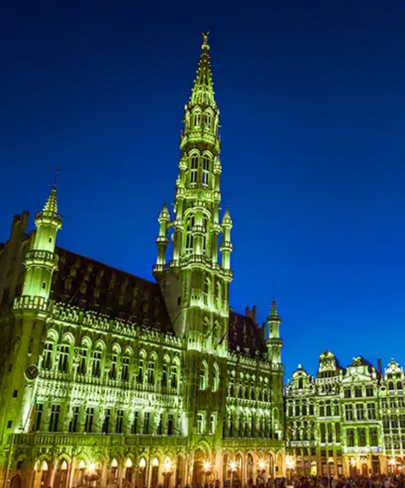 Vue nocturne de l'Hôtel de Ville de Bruxelles illuminé sur la Grand-Place, mettant en valeur son architecture gothique détaillée sur un ciel bleu profond.