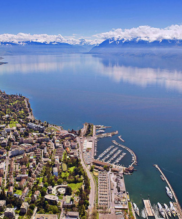 Vue aérienne du quartier d'Ouchy à Lausanne au bord du lac Léman, avec les Alpes françaises se reflétant dans l'eau calme.