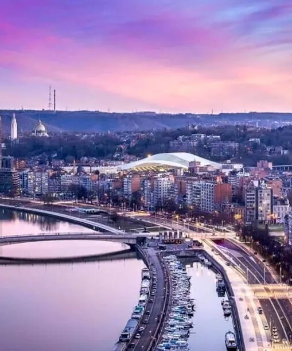 Vue en hauteur de la Meuse à Liège au crépuscule, avec le toit illuminé de la Médiacité et les reflets des lumières de la ville sur l'eau.