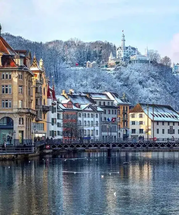 Vue hivernale du front de mer historique de Lucerne, avec des arbres enneigés et un bâtiment aux allures de château sur la colline.