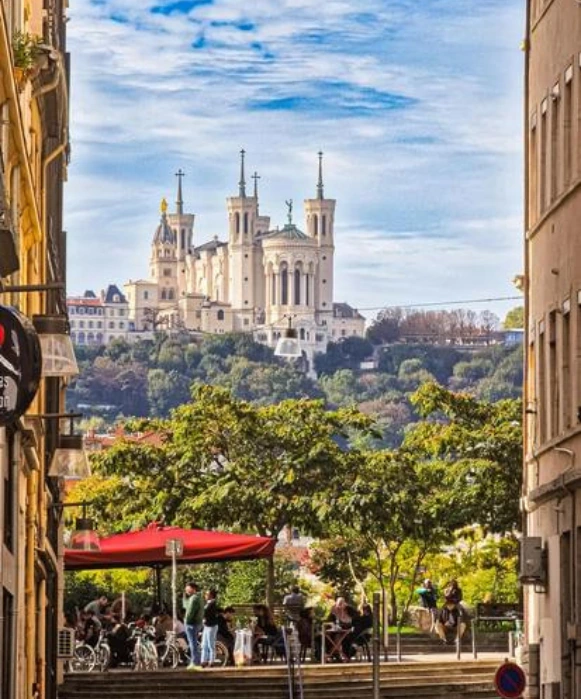 La basilique Notre-Dame de Fourvière sur sa colline, vue depuis une rue charmante du Vieux Lyon avec des gens en terrasse.