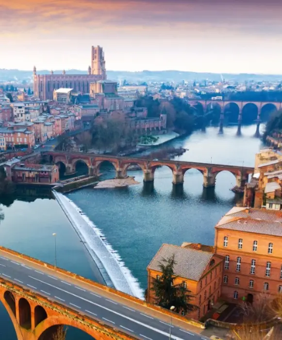 Vue en hauteur d'Albi au coucher du soleil, montrant l'imposante cathédrale Sainte-Cécile en brique, le Tarn et les ponts historiques.