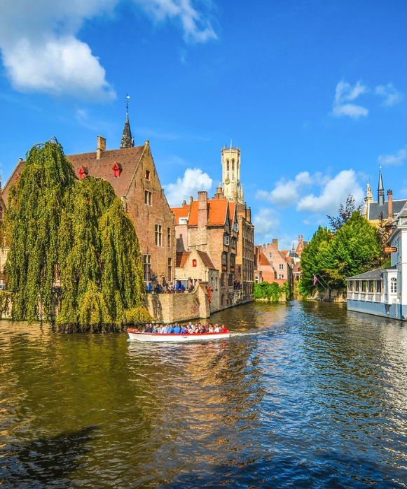 Vue pittoresque d'un canal de Bruges avec un bateau de touristes, des bâtiments historiques en brique et le beffroi en arrière-plan sous un ciel bleu.