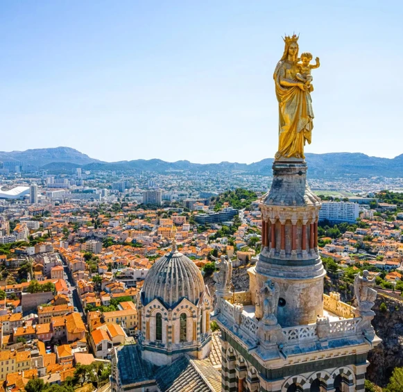 Gros plan sur la statue dorée de la Bonne Mère au sommet de Notre-Dame de la Garde, dominant l'étendue urbaine de Marseille.