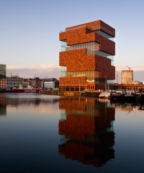 Le musée MAS moderne en grès rouge se reflétant dans les eaux calmes du port au coucher du soleil, avec un navire historique à quai à proximité.