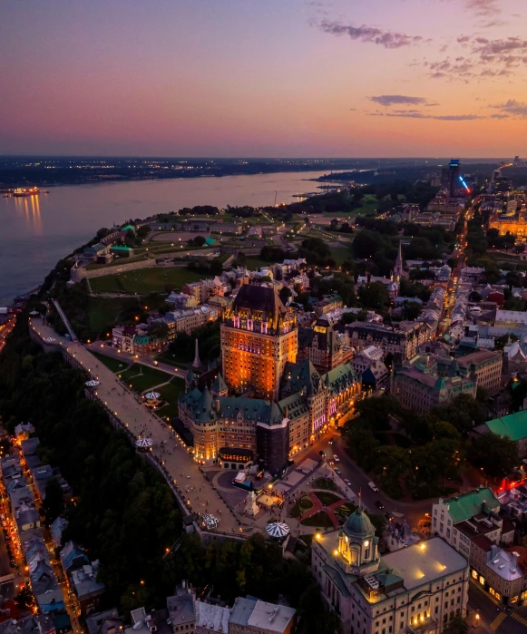 Une vue aérienne spectaculaire du Château Frontenac illuminé et de la terrasse Dufferin au coucher du soleil, avec le fleuve Saint-Laurent et la rive de Lévis en arrière-plan.