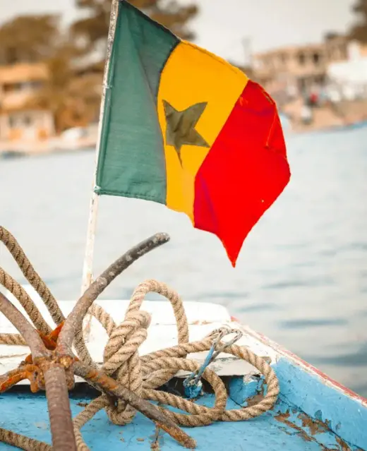 Gros plan sur le drapeau du Sénégal flottant à la proue d'une pirogue traditionnelle, avec une ancre rouillée et des cordages au premier plan sur fond de mer brumeuse.