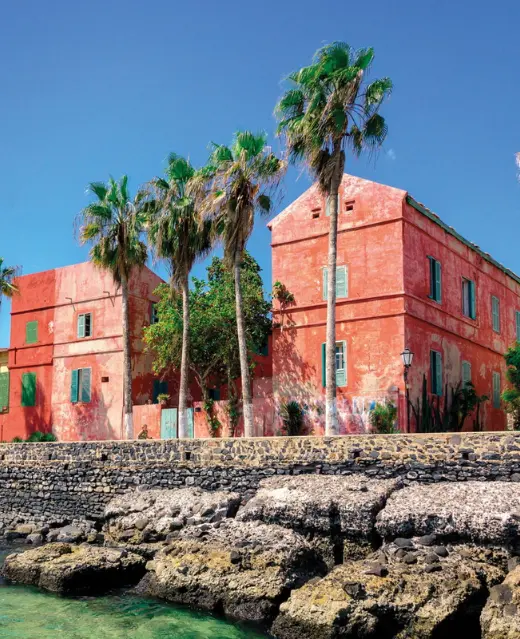 Vue ensoleillée des bâtiments coloniaux rouges emblématiques de l'île de Gorée, encadrés par de hauts palmiers et la côte rocheuse sous un ciel bleu pur.
