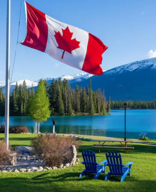 Le drapeau canadien flotte fièrement devant un paysage de montagne serein, avec des chaises Adirondack bleues au bord d'un lac turquoise et des sommets enneigés.
