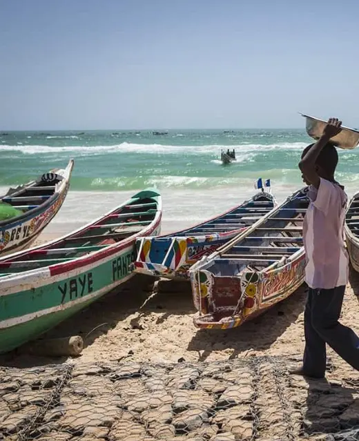 Pirogues sénégalaises traditionnelles et colorées alignées sur la plage de sable de Yoff, tandis qu'un jeune garçon passe devant sous un soleil radieux.