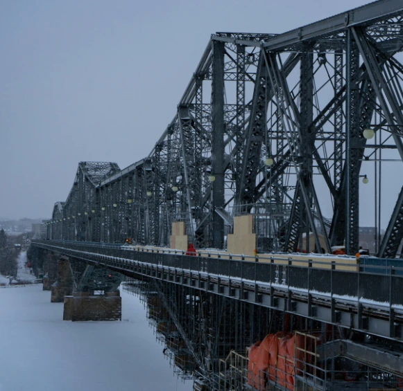 Une vue en perspective de l'imposante structure d'acier du pont Alexandra recouverte de neige par une journée d'hiver grise.