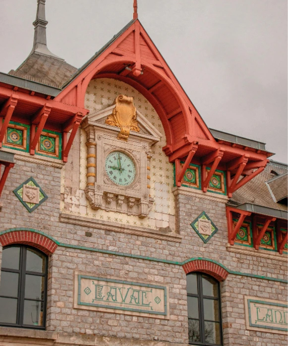 Gros plan sur le bâtiment historique en pierre de la gare de Laval, avec son horloge décorative, ses pignons en bois rouge et ses accents de tuiles colorées.